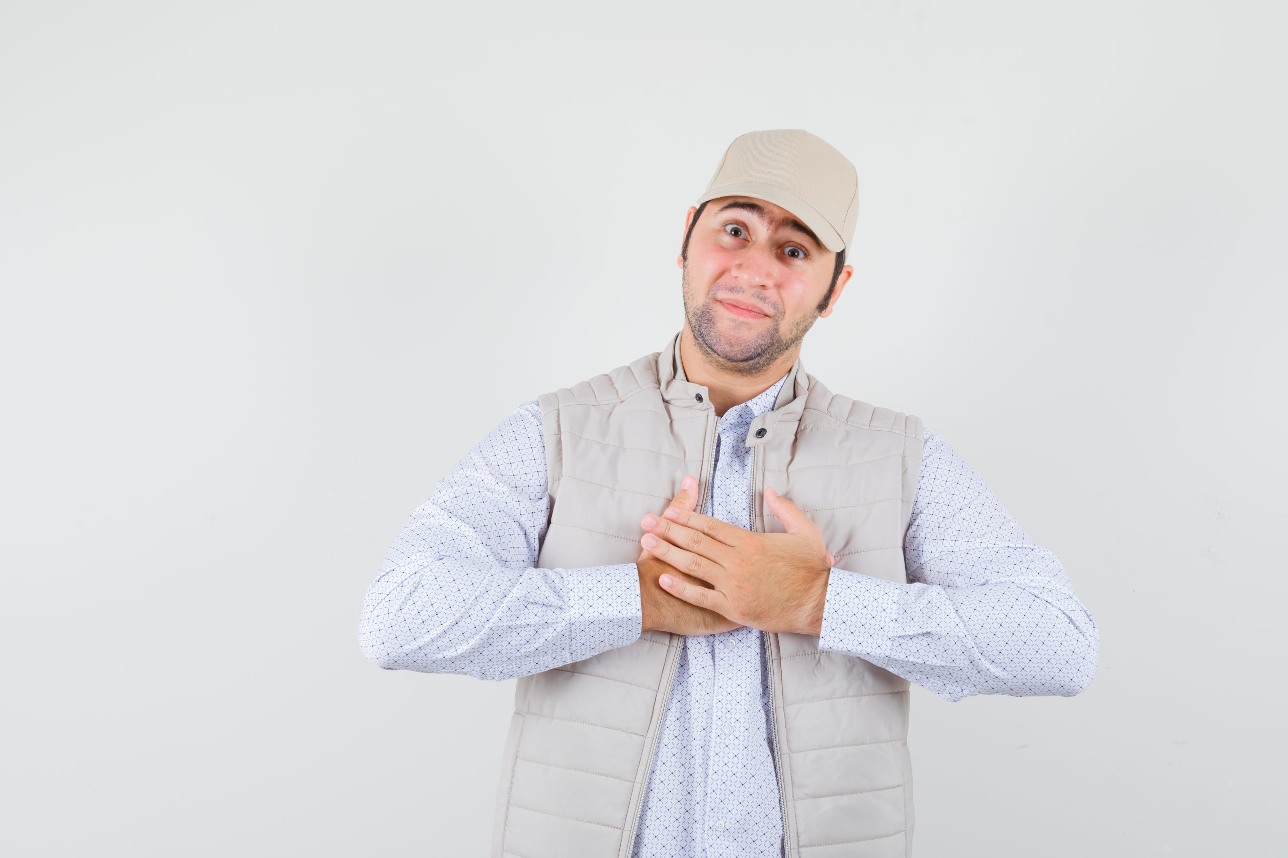 young man in shirt,sleeveless jacket,cap holding hands on his chest and looking thankful , front view.
